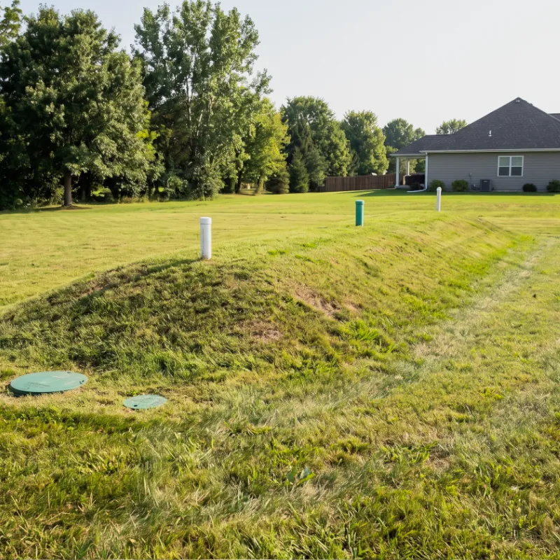 Septic mound system with white inspection pipes on a grassy rise in a country yard, with a home beyond