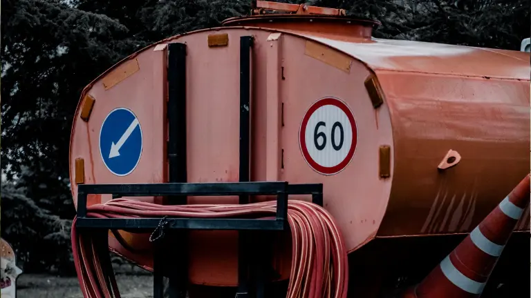 Rear of an orange tank trailer with a coiled red suction hose on its rack and a traffic cone beside it